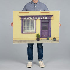 A person from The Canvas Works holding a 100x70cm canvas print of a yellow storefront with a purple door and window, featuring the text 'J O'DONOVAN'.  The storefront is located in Courtmacsherry near Kinsale. 