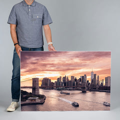 A man in a gray polo shirt and jeans holds a large rectangular 120x80cm canvas print showing the New York City skyline at sunset over the harbor, with boats and bridges in the foreground.