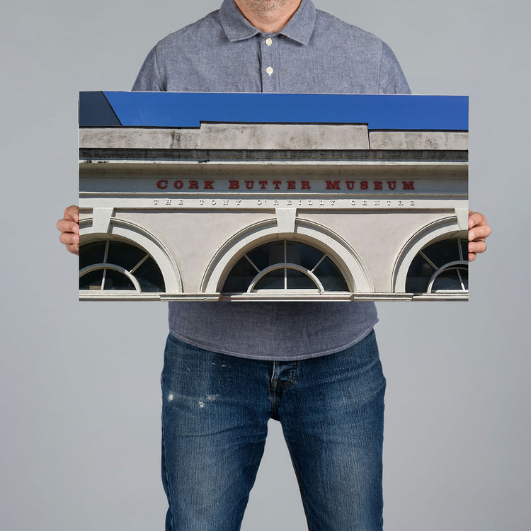 A man holds a 60x30cm panoramic canvas print art featuring the facade of Cork Butter Museum. The print has a blue upper section and a light gray lower section with red lettering.