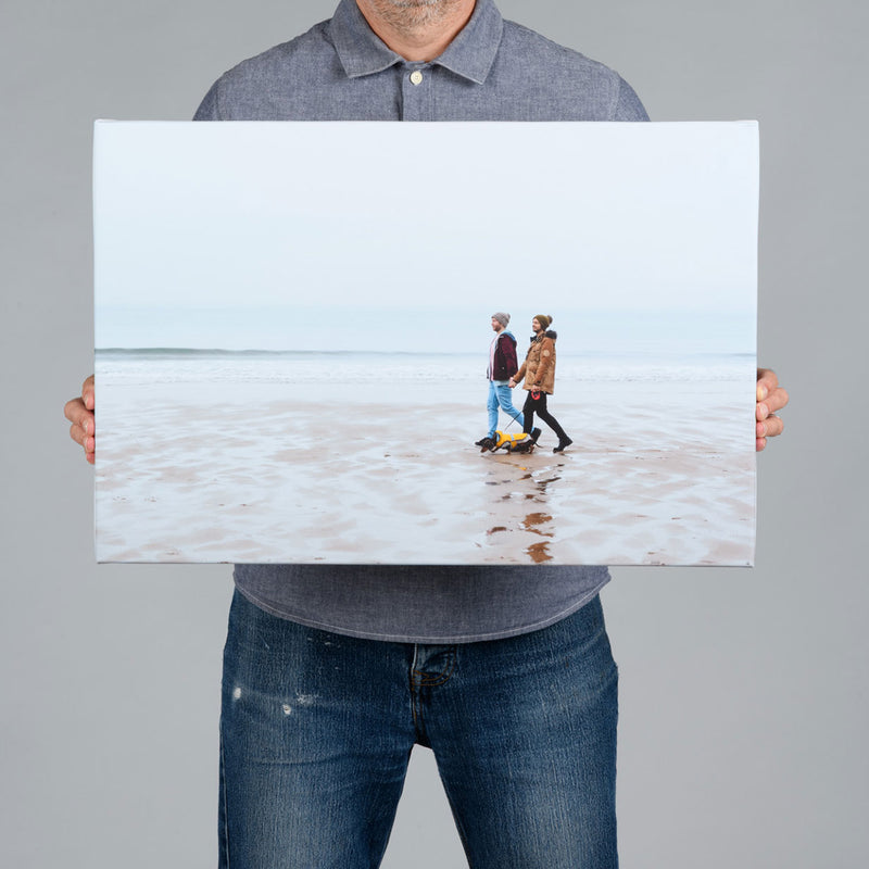 A man holding a 60x40cm canvas print featuring a same sex couple walking on the beach with their dog