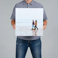 A man from The Canvas Works  holds up a square 40x40cm canvas print showing a beach scene with two people walking on the sand.