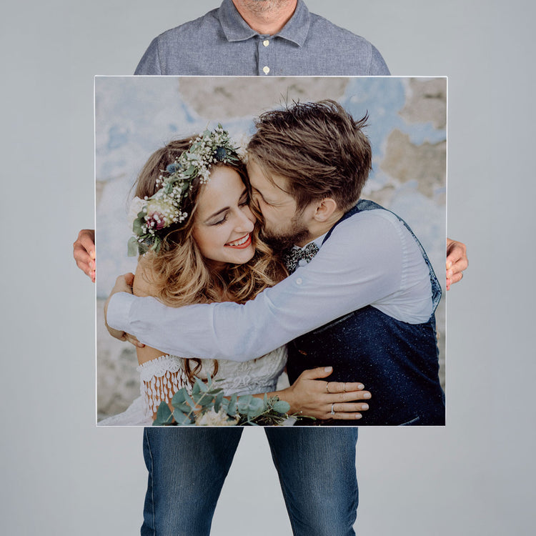 A man from The Canvas Works holds up a square 60x60cm canvas print featuring a bride and groom hugging. 