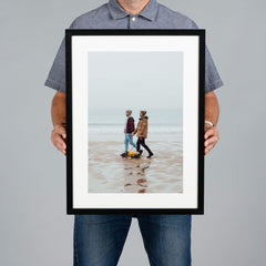 A man holds up a 30x45cm black framed art print against a solid gray background. The print features a photograph of a couple walking on a beach, framed with a white mat and a black frame.