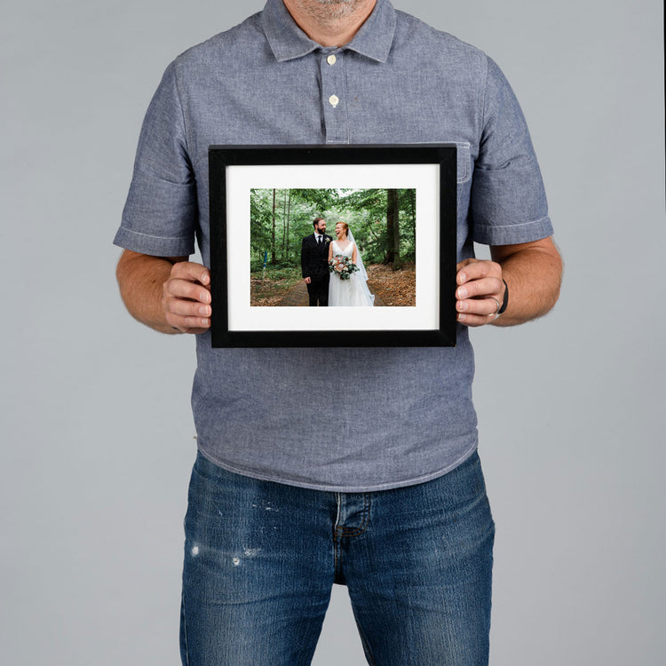 A man holds a 20x15cm framed photo print against a solid gray background. The frame is black with a white mat, showcasing a custom wedding photograph of a couple in a forest.