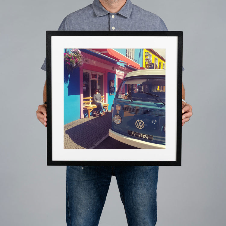 A person holding a 40x40cm black framed art print featuring a colorful street scene with a blue Volkswagen van parked outside a bright blue shop. 