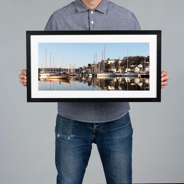 A person holds a black framed art print of a harbour scene in Kinsale with boats and masts against a plain gray background. The frame is black witha white matt. 