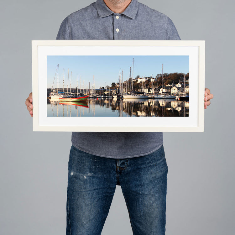 A person holds a white framed art print of a harbour scene in Kinsale with boats and masts against a plain gray background. The frame is white with a white matt. 