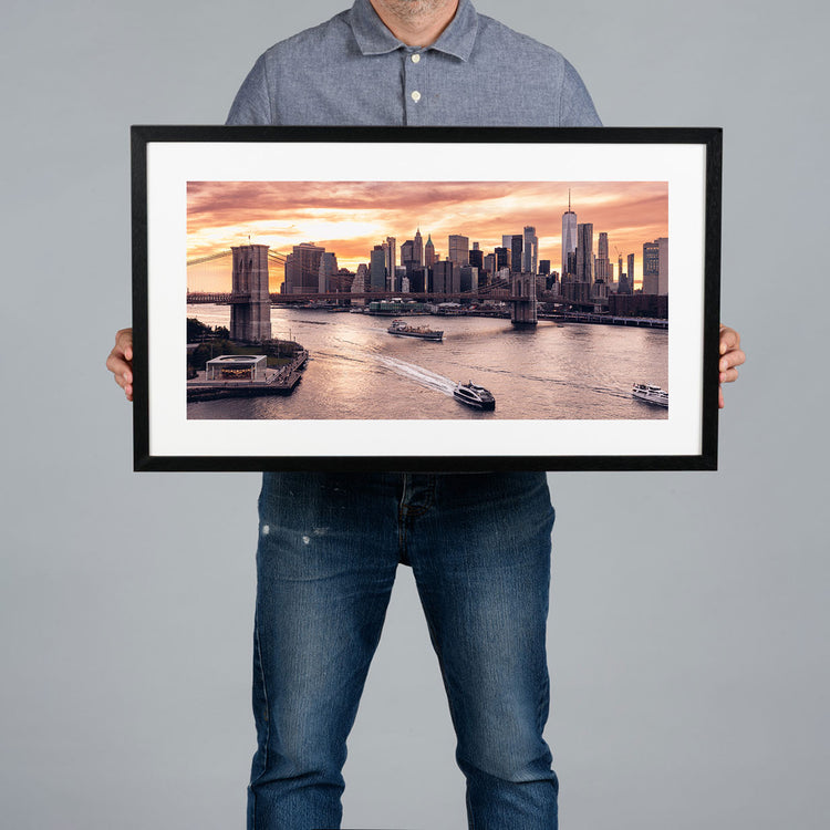 A person holding a 60x30cm framed panoramic print of a cityscape of the New York City skyline at sunset.