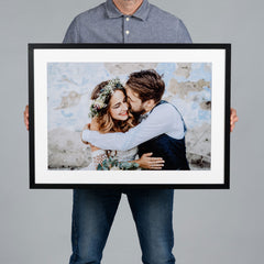 A man holds a large 60x40cm black-framed custom photo print featuring a wedding couple embracing. The print has a wide white mat border and is displayed against a solid gray background.